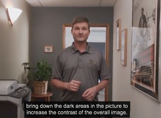 A man in a grey polo shirt stands in a hallway with a plant and a printer nearby.