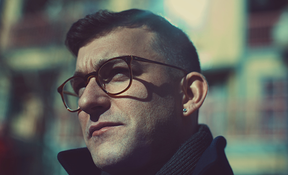 A man with short dark hair and brown glasses looking into the distance outdoors.