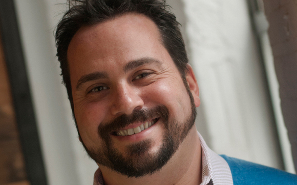 A close-up portrait of a smiling man with dark hair and a groomed beard.