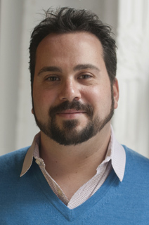 A man with dark hair and a beard wearing a blue sweater over a collared shirt.