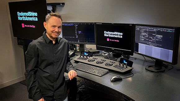 A man in a black shirt stands beside a desk with multiple computer monitors and editing consoles.