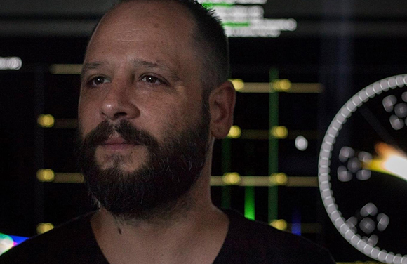 Close-up portrait of a man with a dark beard and short hair looking upward against a dark, blurred background.