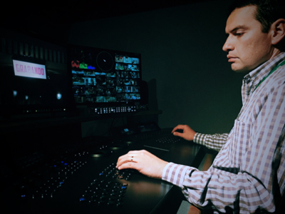 A man in a checkered shirt operates a control panel in a dark room with multiple video monitors.