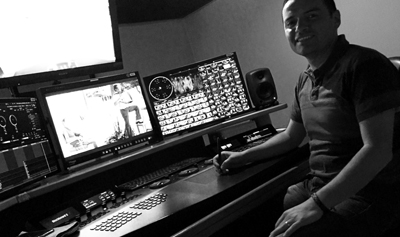 A man smiles while sitting at a desk with multiple computer monitors and a large control console.