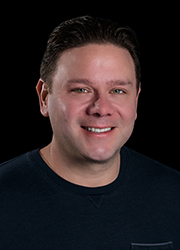 A smiling man with short dark hair wearing a dark crewneck shirt against a black background.