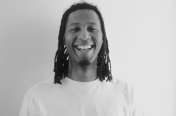 A black and white portrait of a man with dreadlocks smiling at the camera.