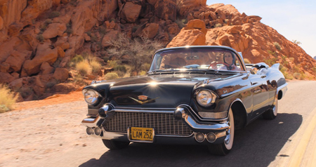 A black vintage convertible drives along a paved road through red rock desert formations.