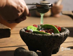 A hand uses a metal juicer to squeeze lime juice into a stone bowl containing guacamole and small dice.