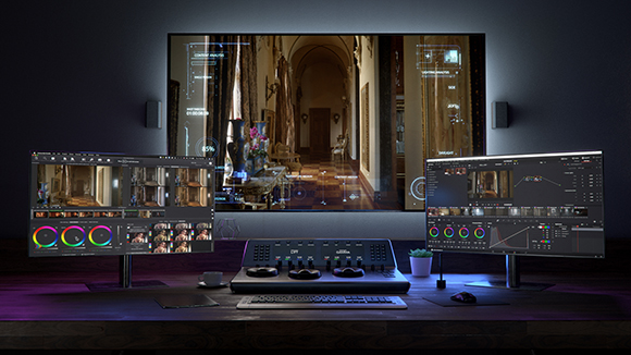 Three computer monitors displaying video editing software and a control surface on a dark wooden desk.
