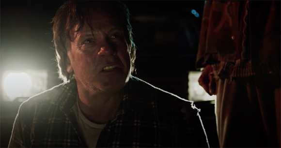 A man in a plaid shirt looks upward with a worried expression in a dimly lit setting.
