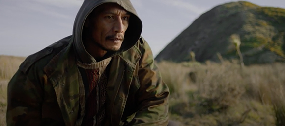 A man wearing a grey hood and camouflage jacket looks into the distance in a grassy field.