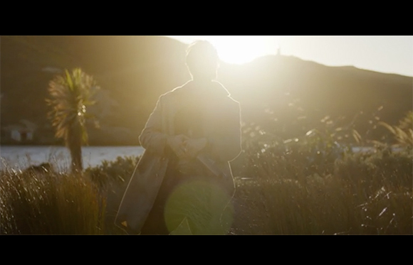 A person in a light coat stands in a field facing a bright sunset over hills.