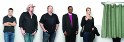 Six diverse people stand in a line against a white backdrop next to a green curtain.