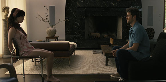 A woman in a pink dress and a man in a blue shirt sit facing each other in a modern living room.
