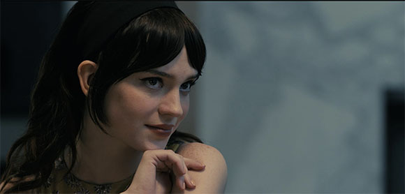 A woman with dark hair and bangs rests her chin on her hand while looking off-camera.