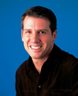 A smiling man with short dark hair wearing a dark button-down shirt against a solid blue background.