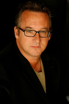A man with short brown hair and glasses wearing a black blazer over a tan shirt.
