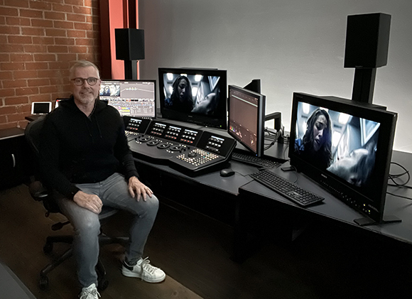 A man sits in a chair before a desk with multiple monitors and a large video editing console.
