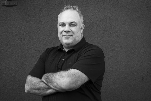 A smiling man with short hair and a black polo shirt stands with his arms crossed against a dark wall.