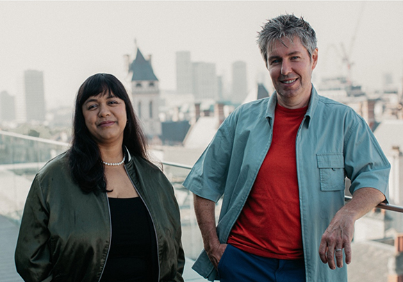 A man in a light blue shirt and red t-shirt and a woman in a dark jacket stand on a rooftop.