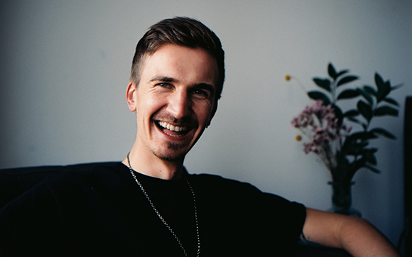 A smiling man with short brown hair and a goatee wearing a black t-shirt and a silver chain.