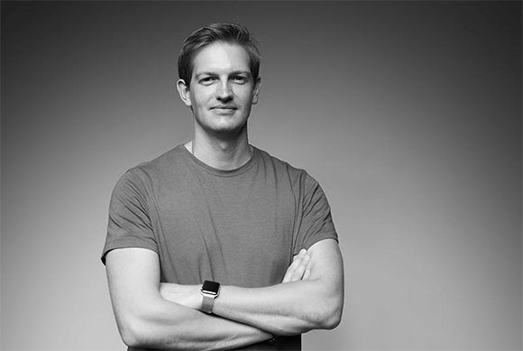 A man with short hair wearing a t-shirt and a smartwatch, posing with his arms crossed against a gray background.