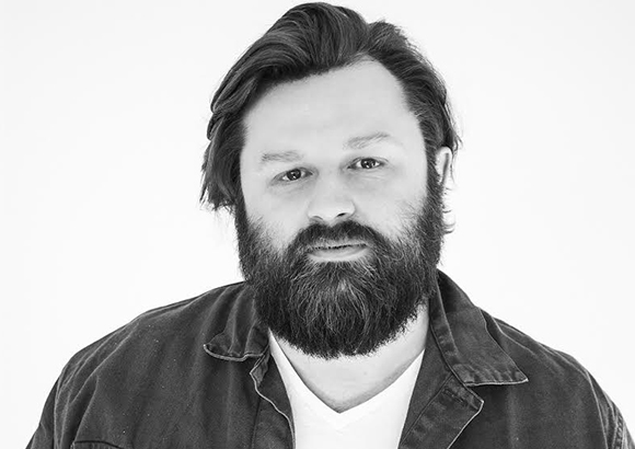 Black and white headshot of a man with dark hair and a full beard wearing a dark jacket over a white shirt.