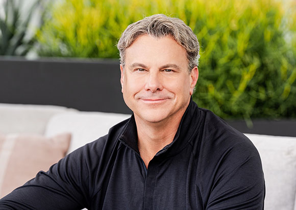 A man with salt-and-pepper hair smiling while wearing a black long-sleeved shirt against a green background.