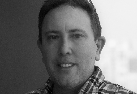 Black and white headshot of a man wearing a plaid button-down shirt looking toward the camera.