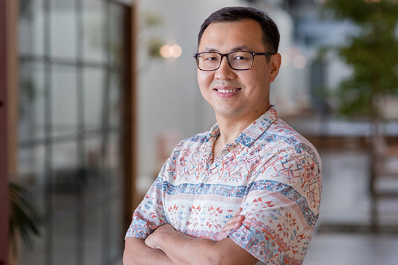 A man with glasses and a patterned short-sleeve shirt smiles with his arms crossed.