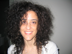 A woman with voluminous, dark curly hair smiling at the camera against a plain wall.