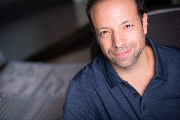 A man with short dark hair and light stubble wearing a blue polo shirt, smiling.