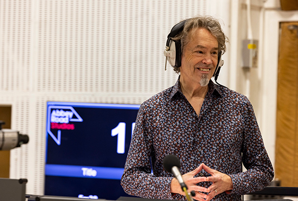 A man with graying hair and a goatee wearing headphones and a patterned shirt in a studio.