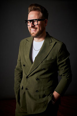 A smiling man with glasses and a beard wearing an olive green double-breasted blazer and a white t-shirt.