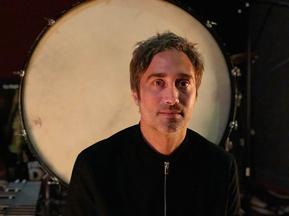 A man with short brown hair wearing a black jacket, positioned in front of a large white drum head.