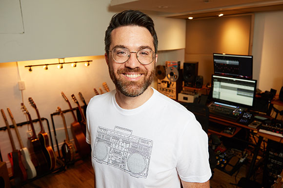 A man with glasses and a beard smiles in a music recording studio with guitars and equipment.