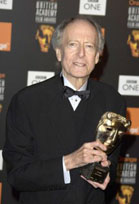 An older man in a black tuxedo and bowtie holds a gold mask-shaped award against a branded backdrop.