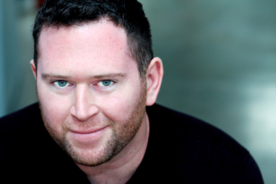 A man with short dark hair, blue eyes, and a light beard looking toward the camera.