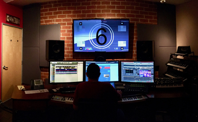 A person sits at a desk with multiple computer monitors and studio equipment in a room with a brick wall.