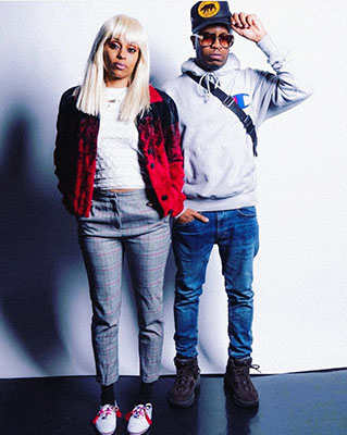 A blonde woman in a red jacket and plaid pants stands next to a man in a grey hoodie and blue jeans.