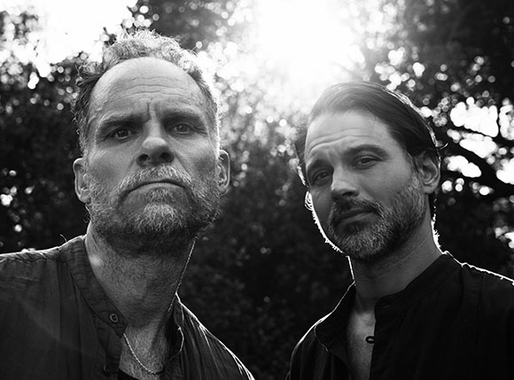 Black and white portrait of two men with facial hair looking at the camera against a backdrop of trees and sunlight.