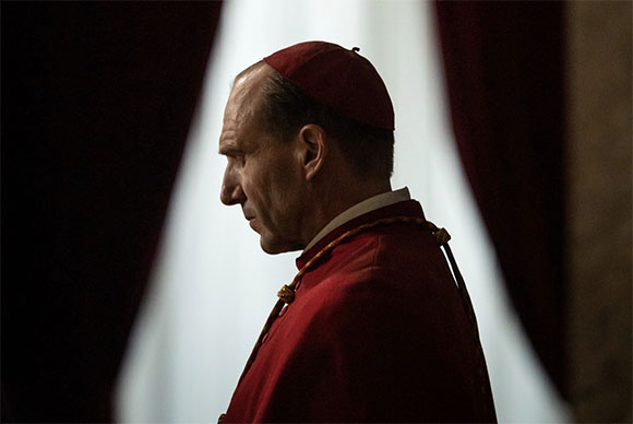 Profile of a man wearing a red zucchetto and red clerical robes against a backdrop of white and red curtains.