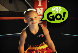 A smiling young boy in a black tank top and boxing shorts inside a boxing ring.