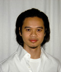 A man with dark, shoulder-length hair and a goatee wearing a white button-down shirt against a white background.
