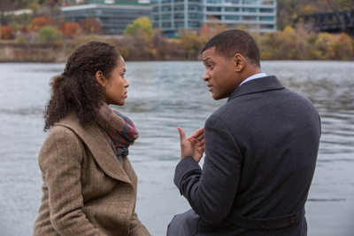 A man in a dark coat and a woman in a brown coat look at each other by a river.