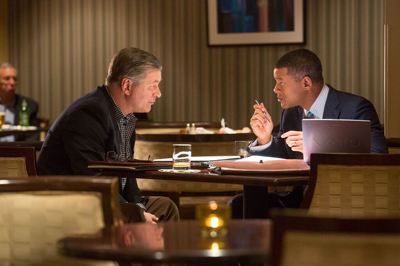 Two men in suits sit across from each other at a dark wooden table in a dimly lit restaurant.