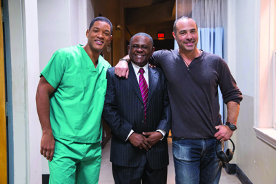 Three men posing together in a hallway, one in green scrubs, one in a suit, and one in a dark long-sleeve shirt.