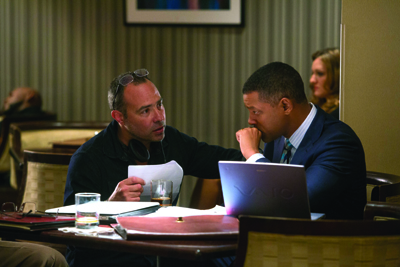 Two men sit at a table in a restaurant, one leaning in to speak while the other looks on near a laptop.