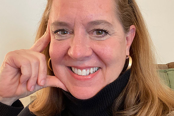 A smiling woman with light brown hair and gold hoop earrings resting her chin on her hand.