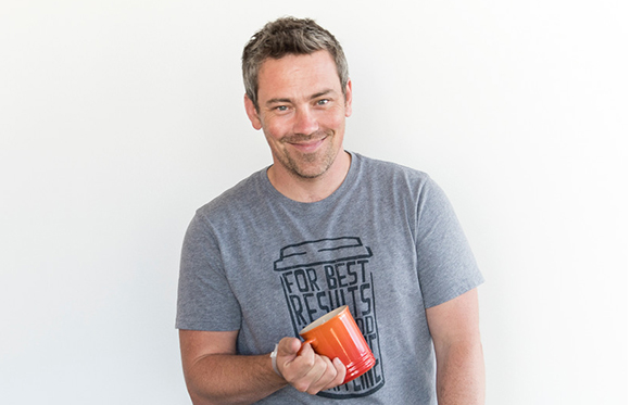 A smiling man in a grey graphic t-shirt holds an orange mug against a white background.